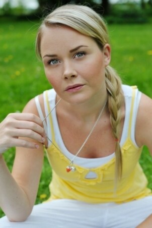 Blond girl wearing a yellow tank top and a silver pendant necklace sitting on the grass